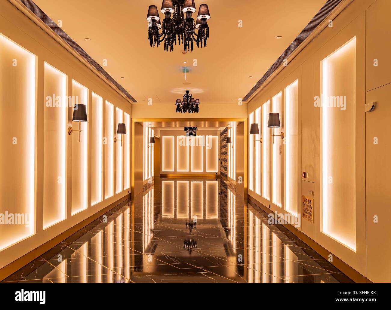 Corridoio lungo con pavimento in marmo scuro, pannelli murali dorati con illuminazione soft e lampadari neri ornati sospesi illuminano il lussuoso Foto Stock