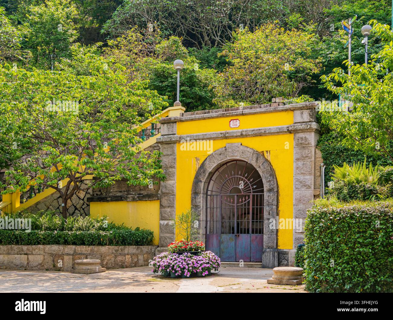 Un arco in pietra con un cancello di metallo e pareti gialle si erge tra una vegetazione rigogliosa con vasi di fiori nelle vicinanze. Foto Stock