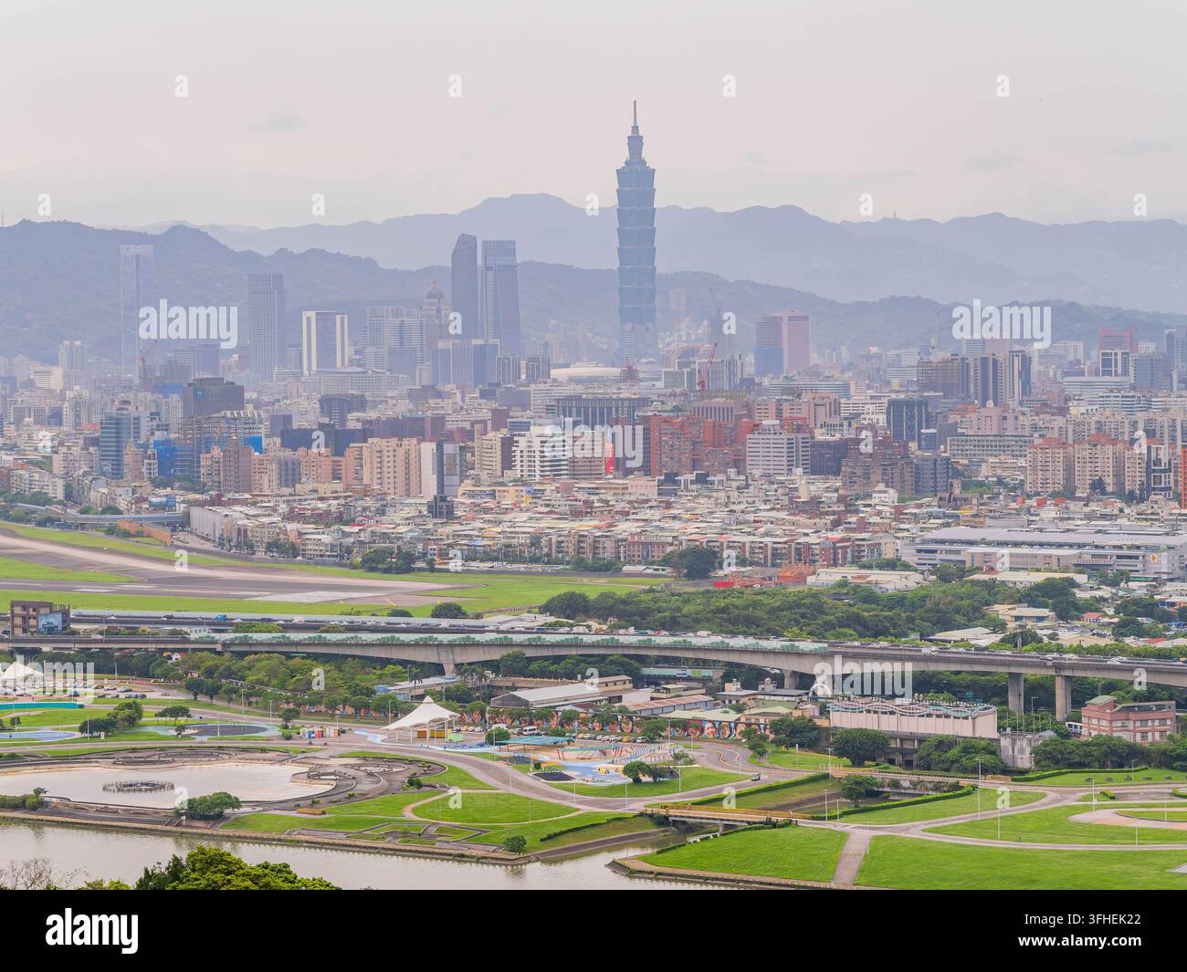 L'iconica torre Taipei 101 si erge alta sulla città, mentre parchi verdi e un fiume riempiono lo sfondo sotto un cielo morbido. Foto Stock