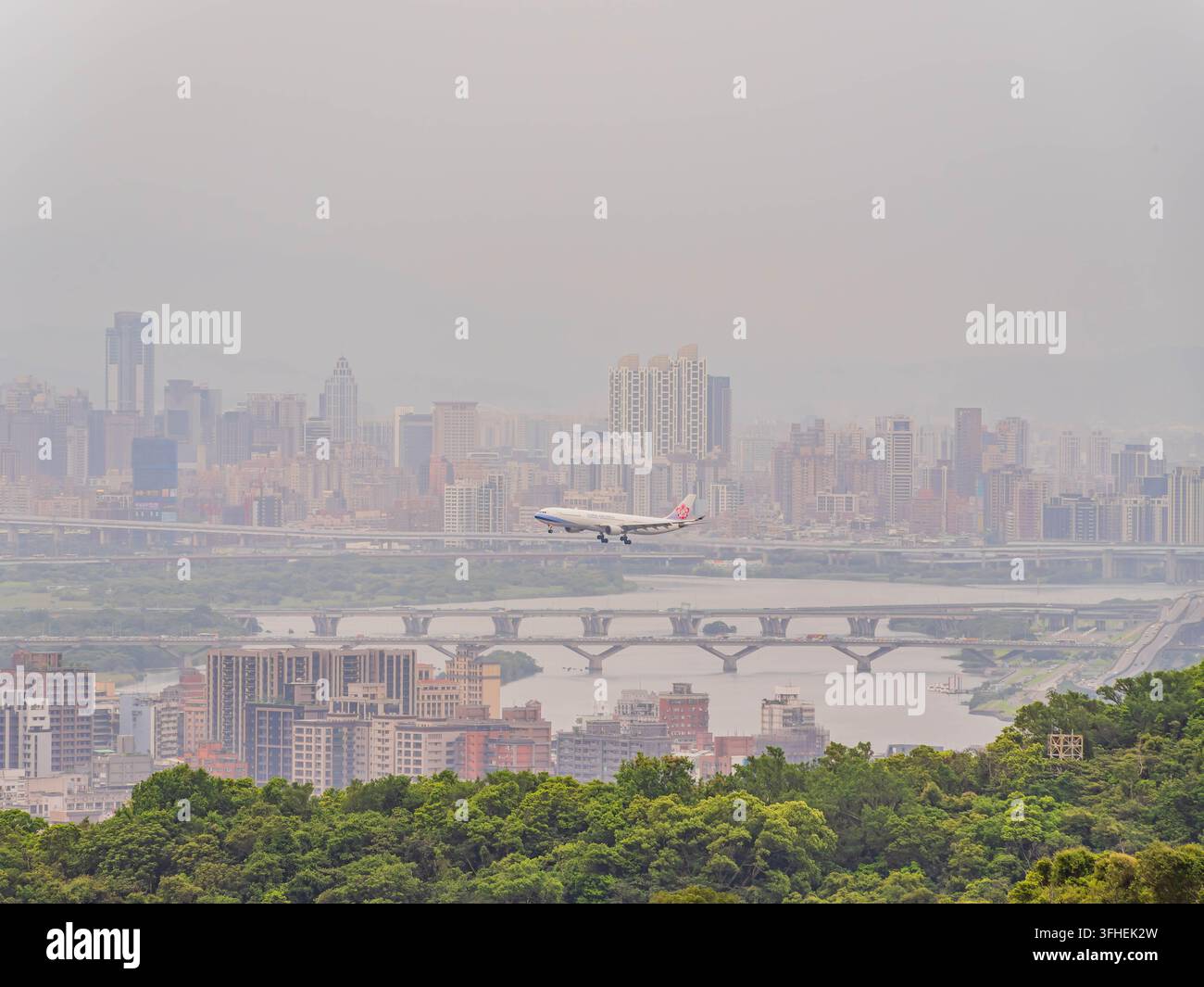 Un aereo scende per l'atterraggio sopra un ampio fiume e un paesaggio urbano sotto un cielo grigio e nebbioso. Foto Stock