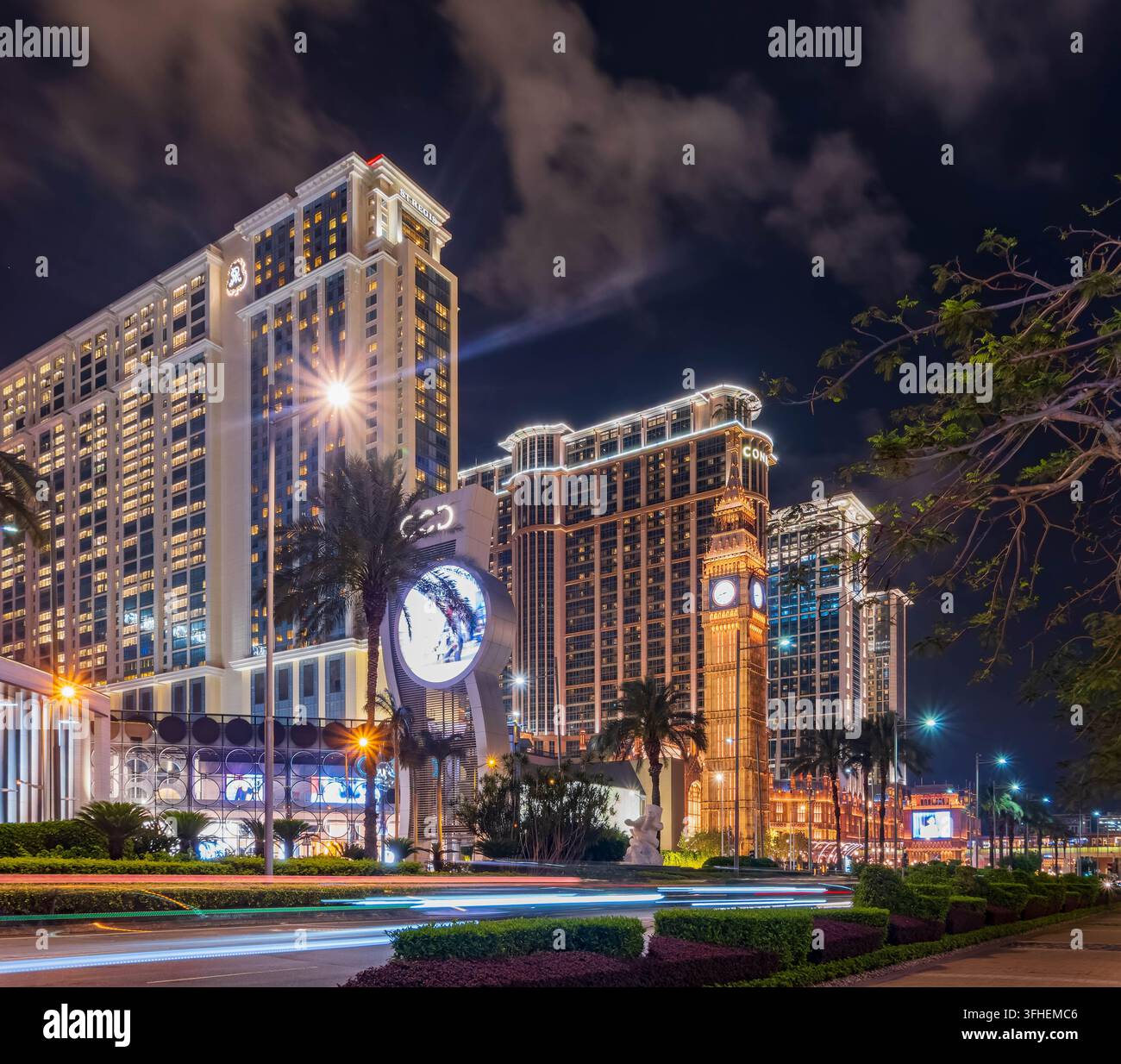 Torri d'albergo illuminati e una torre dell'orologio si ergono dietro una strada con scie di luce dei veicoli in movimento. Foto Stock