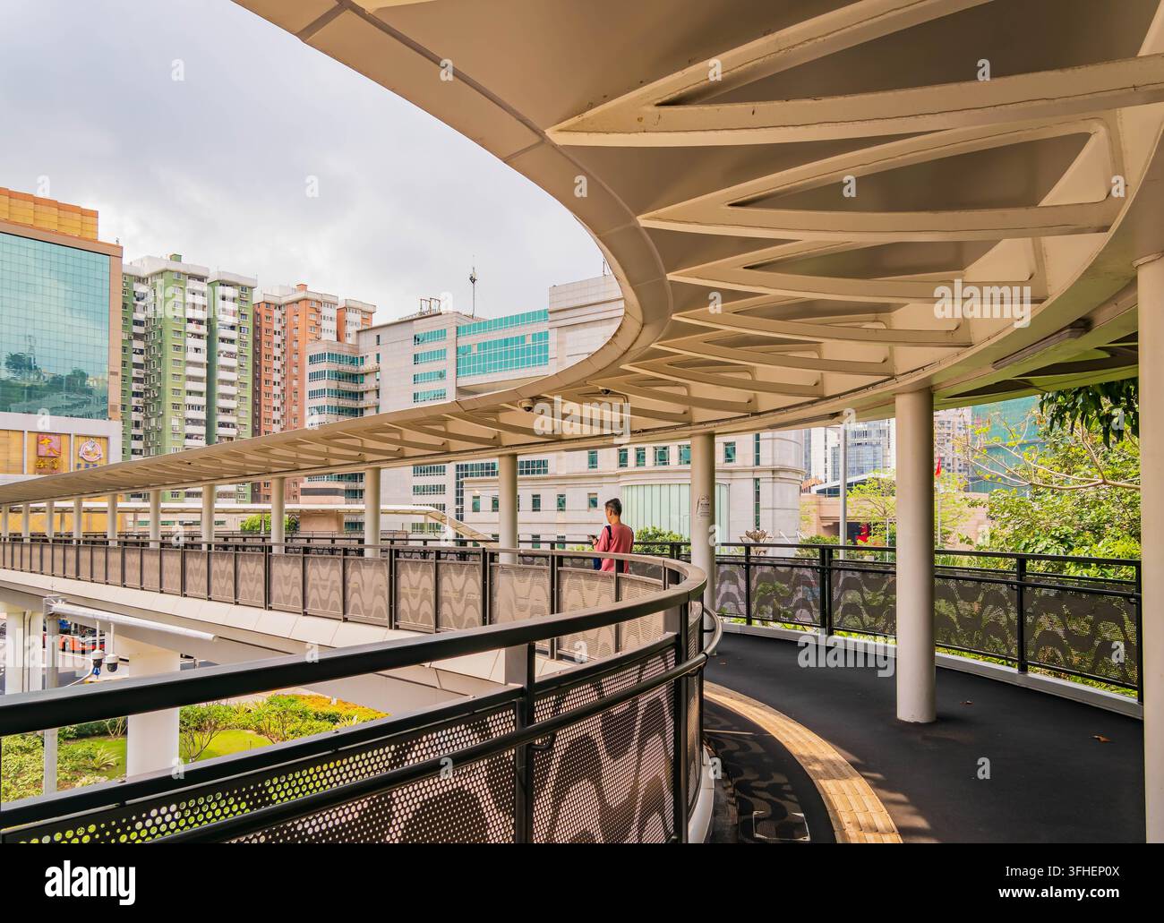 Macao, Cina, 16 APR 2024 – Una figura solitaria passeggia lungo un sentiero nero sotto un baldacchino bianco curvo su un sovrappasso urbano con uno sfondo di città Foto Stock