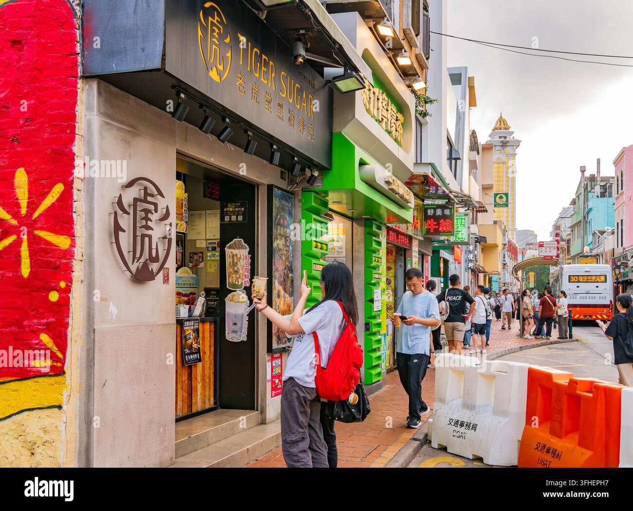 Macao, Cina, 17 apr. 2024 – Una donna tiene il suo tè al boba vicino alla segnaletica del negozio mentre cammina lungo una strada fiancheggiata da negozi colorati e altri Foto Stock