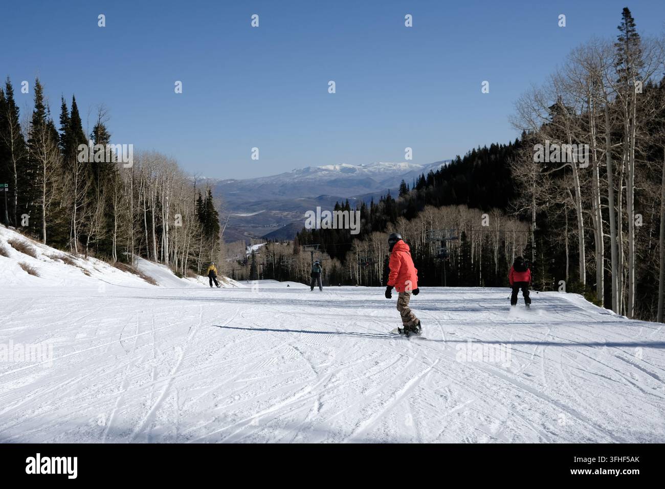 Snowboarder che scendono una pista da sci soleggiata con vista sulle montagne. Giornata invernale soleggiata in una stazione sciistica dello Utah con cime innevate in lontananza. Foto Stock