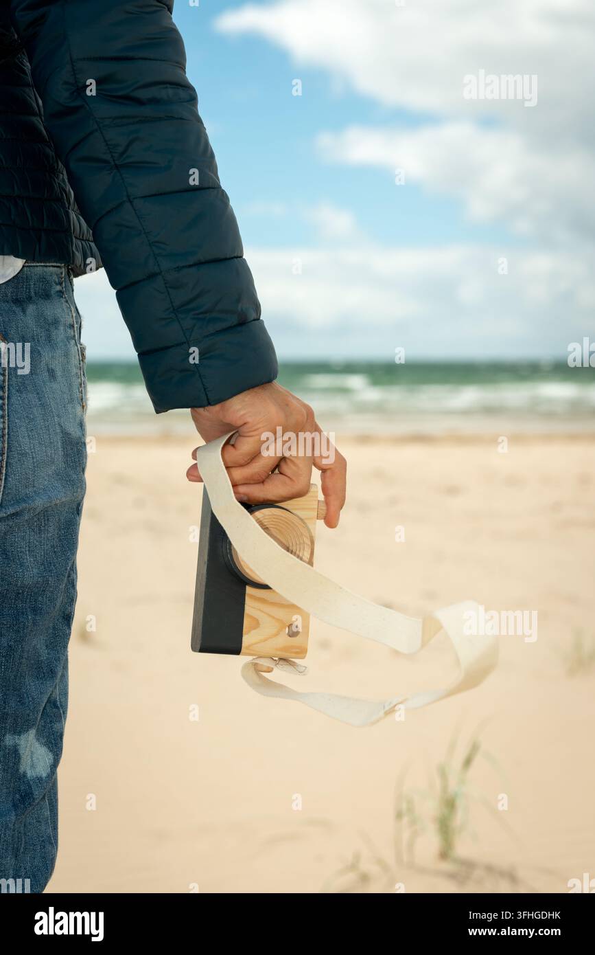 Primo piano della mano di una donna che tiene una fotocamera giocattolo in legno su uno sfondo di spiaggia sabbiosa. Fotografia divertente, concetto di libertà estiva senza fine. Foto Stock