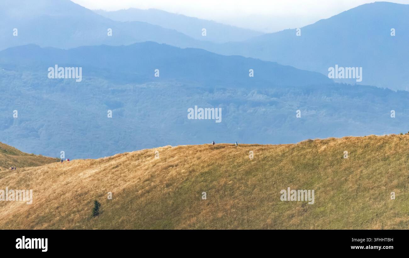 Escursionisti lontani che attraversano un alto crinale erboso sopra valli montane stratificate Foto Stock
