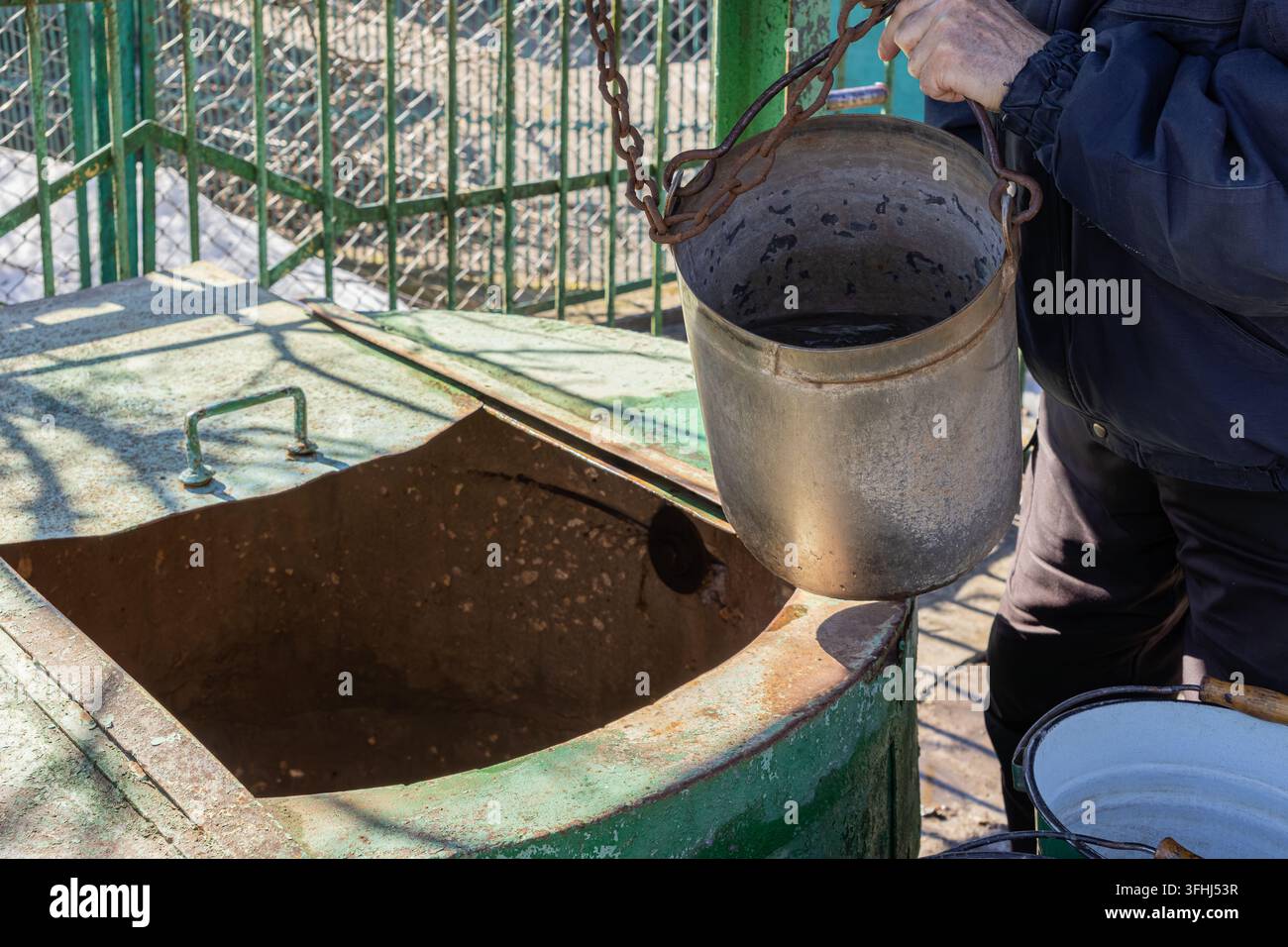 Un individuo tira un secchio di acciaio pesante da un pozzo da giardino stagionato, sottolineando la routine quotidiana della vita di villaggio e la gestione delle risorse naturali con Foto Stock