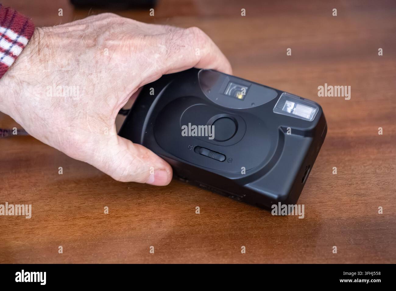 Una mano invecchiata afferra una piccola fotocamera nera punt-and-shoot su una scrivania lucida, rappresentando una passione per la storia fotografica tradizionale e la prese Foto Stock
