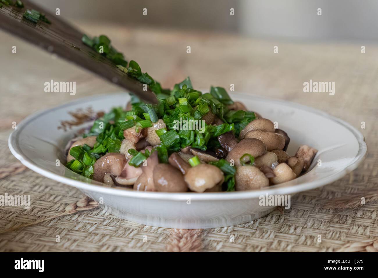 Ciuffo di cipolla primaverile cade da una lama metallica su piccoli funghi cotti all'interno di una ciotola rustica, dimostrando un processo culinario per un pasto nutriente Foto Stock