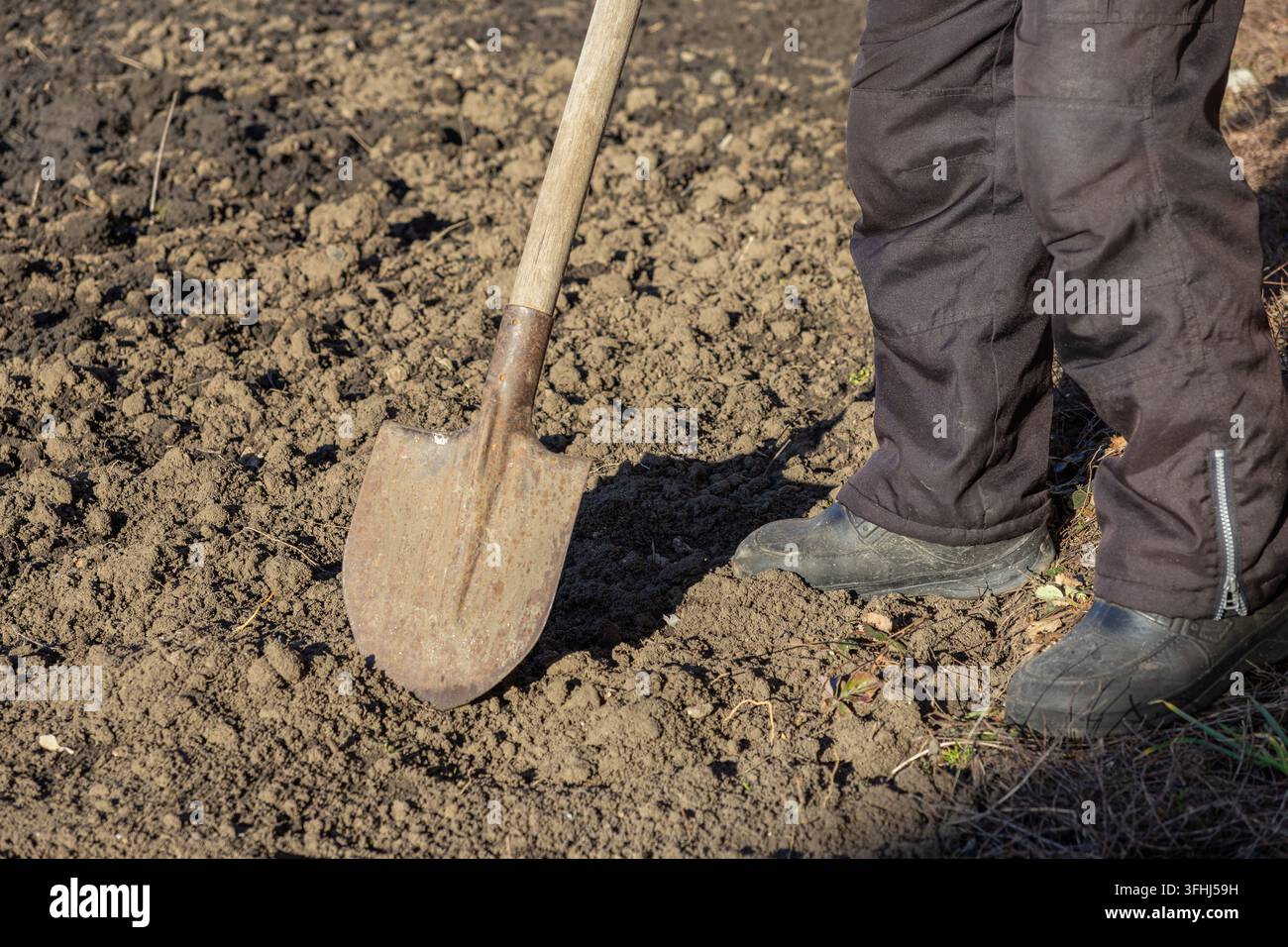Persona con pantaloni scuri in piedi su terra arata che tiene una vanga consumata, illustrando l'attività radicata nel suolo dell'agricoltura e la dedizione necessaria per la produzione Foto Stock