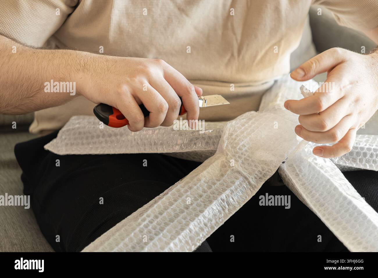La persona taglia con cura il materiale di imballaggio in plastica con una lama affilata, catturando il compito pratico di disimballare beni acquistati tramite e-commerce Foto Stock