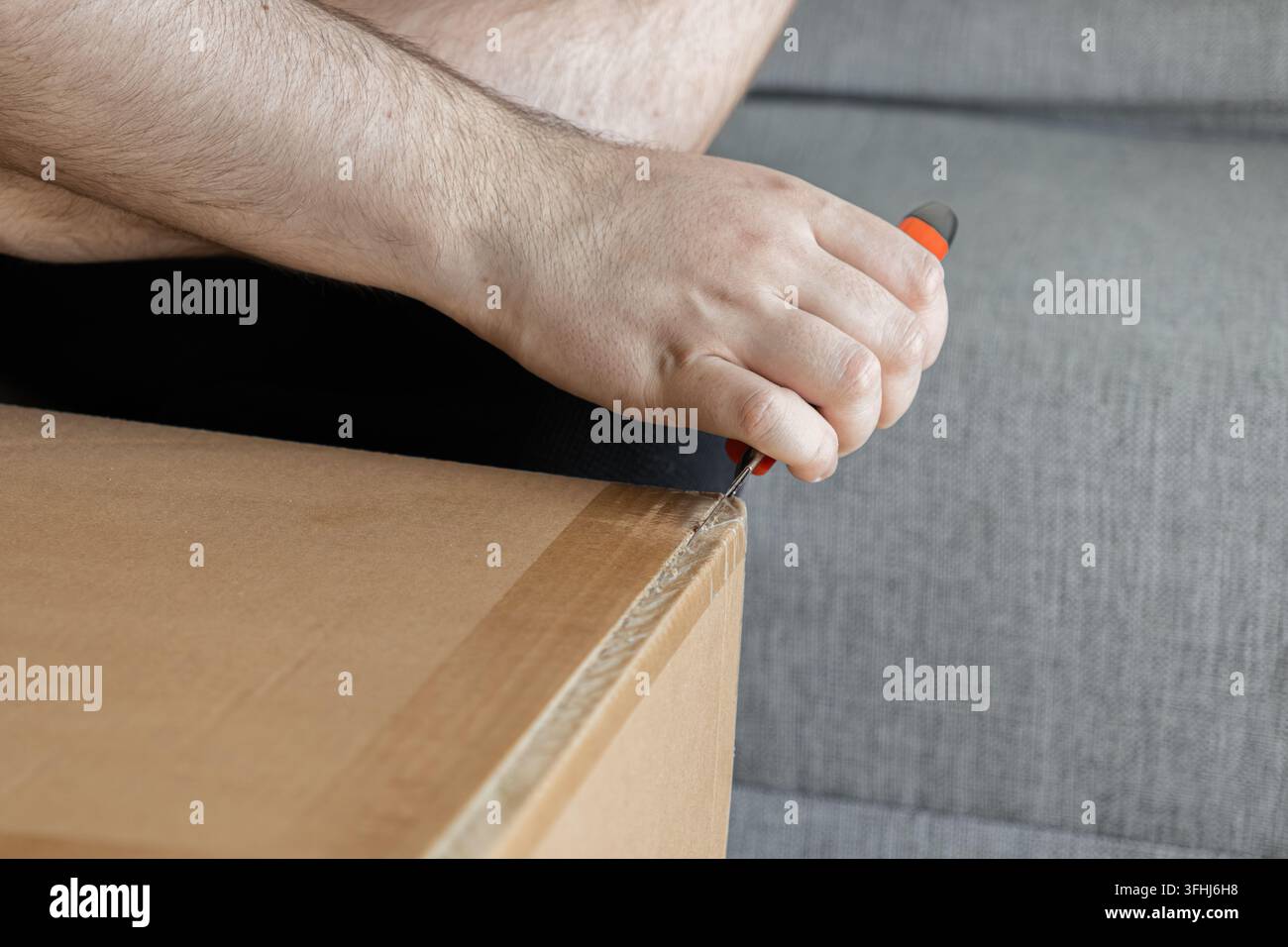 Una persona usa un taglierino affilato per tagliare il nastro adesivo su un cartone da spedizione marrone, illustrando l'eccitazione di aprire un ordine online all'interno di una c Foto Stock