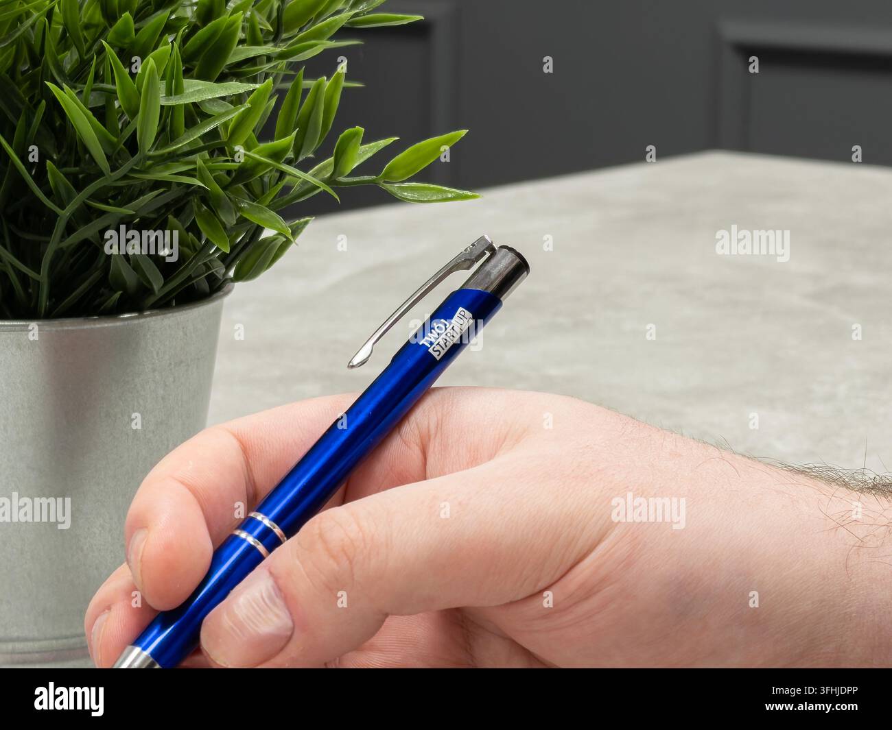Poznań, Polonia - 16 gennaio 2026: Mano che tiene una penna a sfera blu brillante vicino a una pianta da scrivania, rappresentante la cancelleria aziendale, la scrittura professionale, Foto Stock