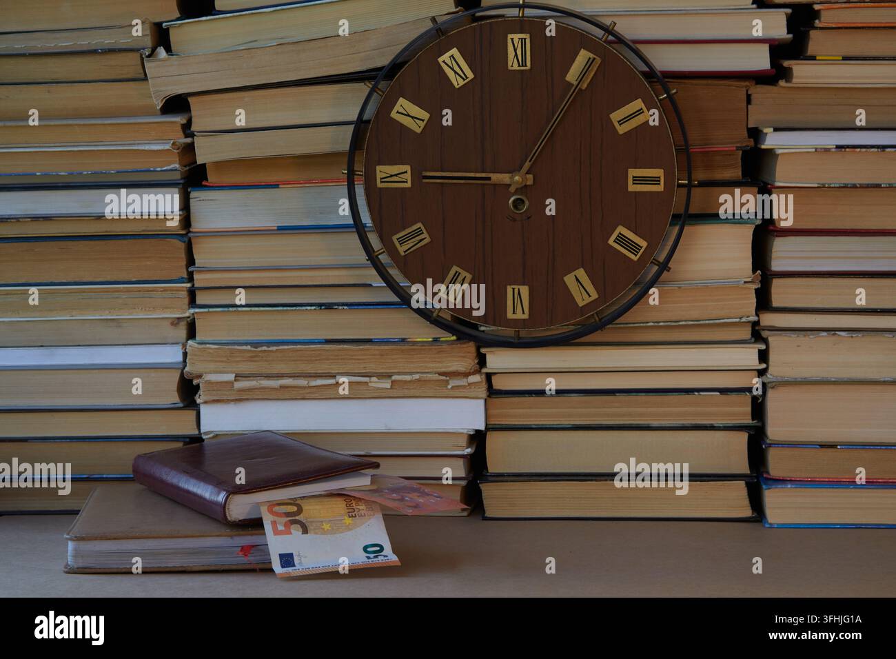 Grande orologio con numeri romani sul quadrante contro uno sfondo di libri impilati e organizzatori posti nelle vicinanze, da cui sono visibili euro. Foto Stock
