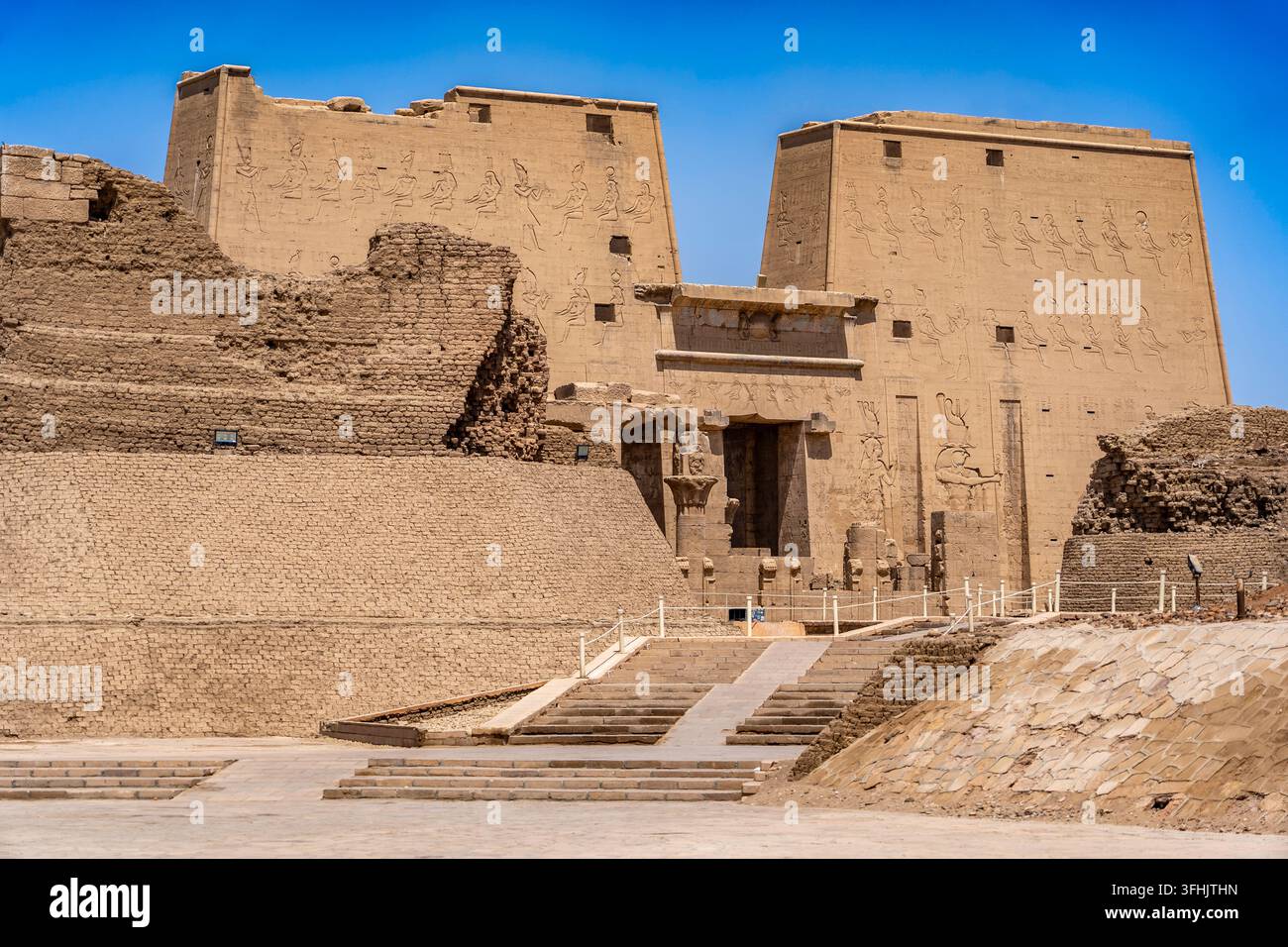 In primo piano, l'imponente pilone d'ingresso del Tempio di Horus a Edfu, circondato dai resti archeologici di un'antica struttura in mattoni di fango Foto Stock