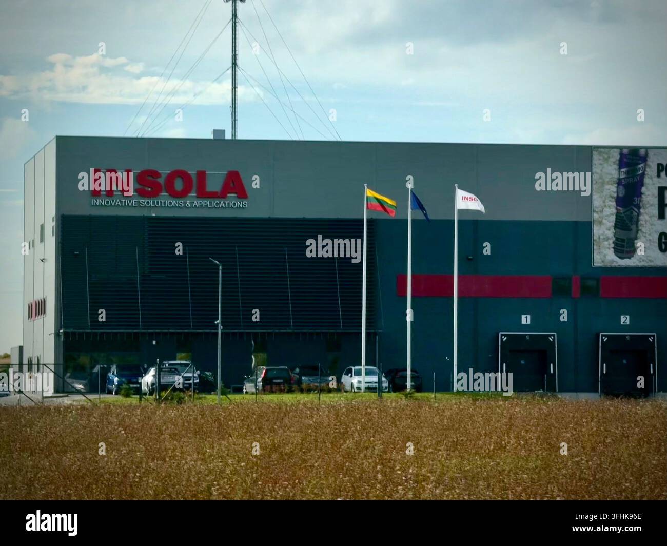 Struttura moderna di ingegneria e soluzioni solari INSOLA in Lituania, con bandiere aziendali e una torre di comunicazione oltre un campo autunnale dorato - Immagine stock catturata con smartphone