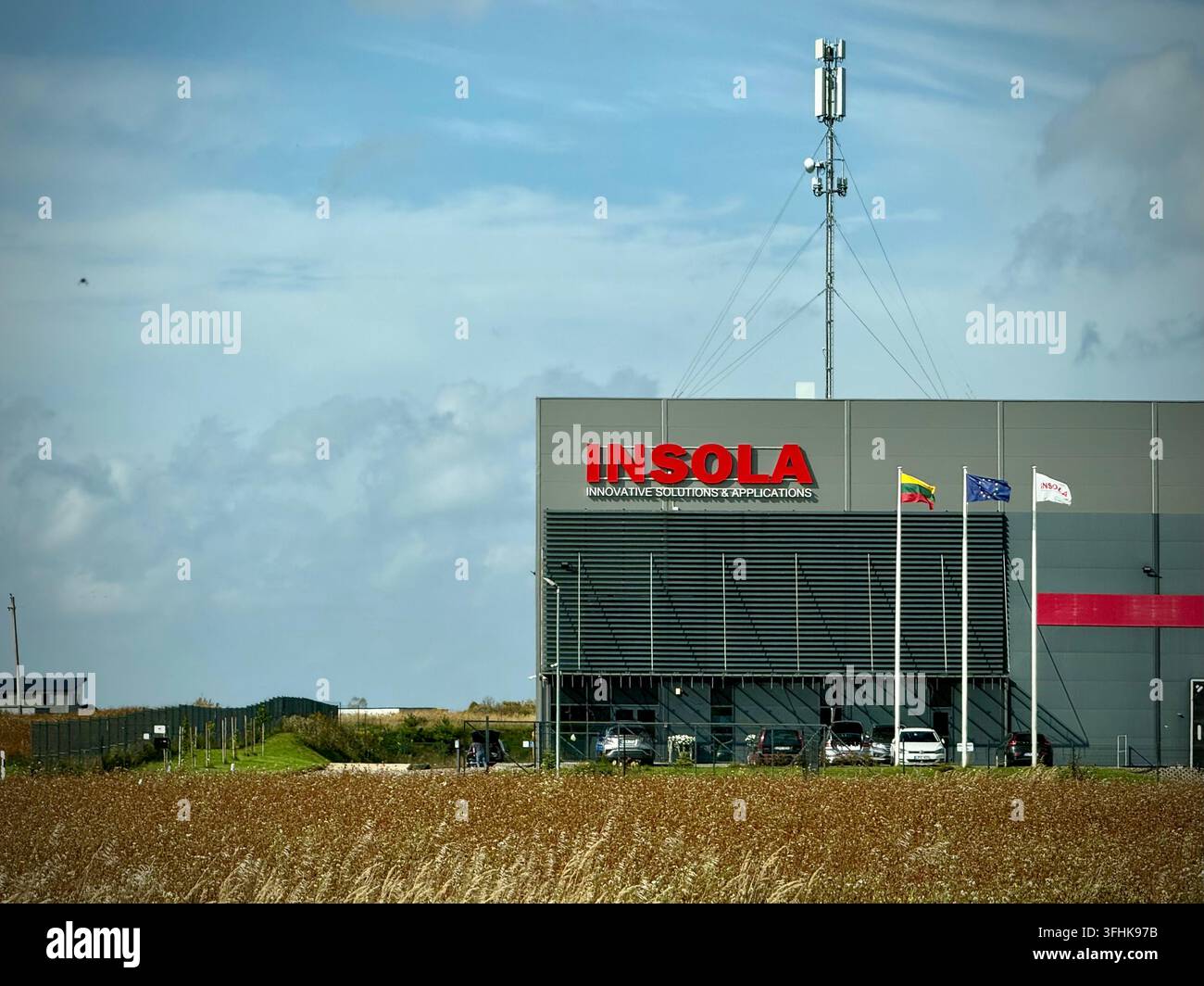 Struttura moderna di ingegneria e soluzioni solari INSOLA in Lituania, con bandiere aziendali e una torre di comunicazione oltre un campo autunnale dorato - Immagine stock catturata con smartphone