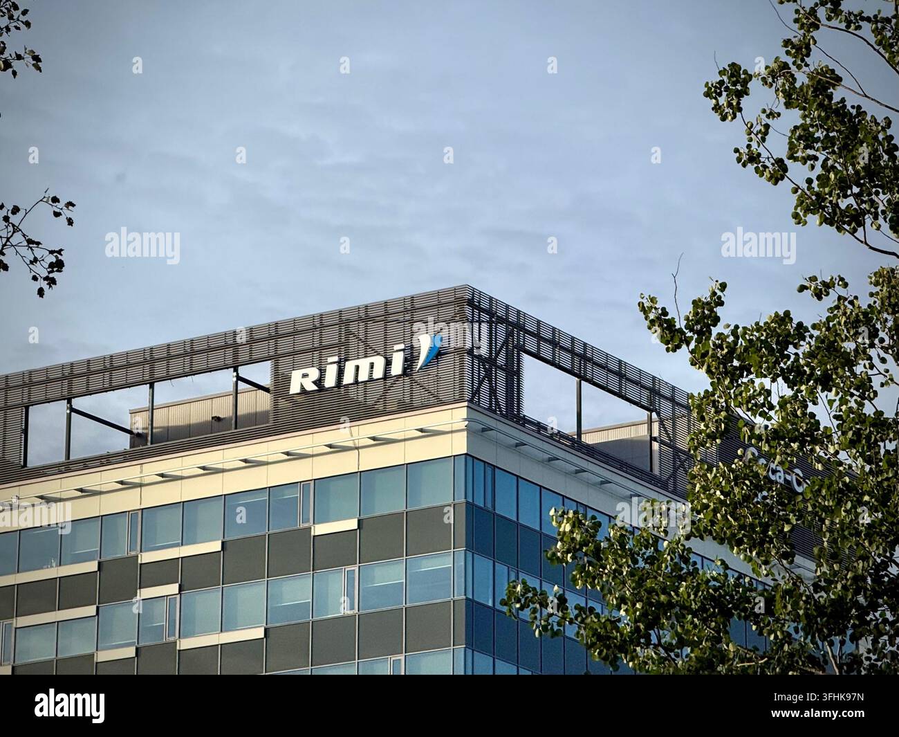 Edificio moderno di ufficio aziendale con il logo della catena di supermercati Rimi sulla linea del tetto, incorniciato dal fogliame verde degli alberi sotto un cielo illuminato dalla luce del giorno. - Immagine stock catturata con smartphone