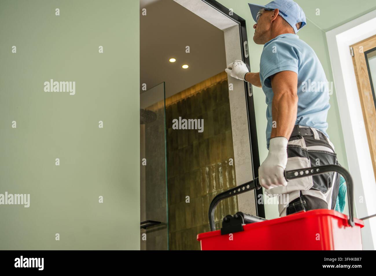Un operaio che indossa guanti e occhiali di sicurezza controlla una cornice della porta mentre tiene una cassetta degli attrezzi accanto all'ingresso del bagno. Foto Stock