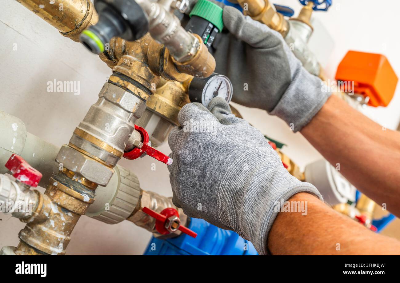 Vista ravvicinata di mani guantate che regolano un regolatore di pressione dell'acqua accanto a un manometro, raccordi per tubi e valvole di intercettazione. Foto Stock
