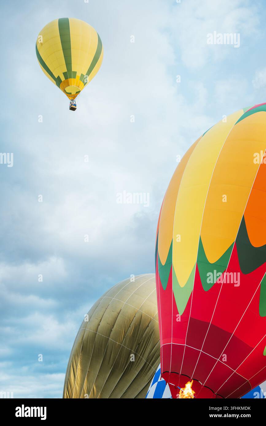 Vibranti palloni aerostatici colorati che si gonfiano con fiamma attiva del bruciatore in primo piano mentre un pallone a strisce gialle vola in alto in un cielo blu nuvoloso durante o Foto Stock