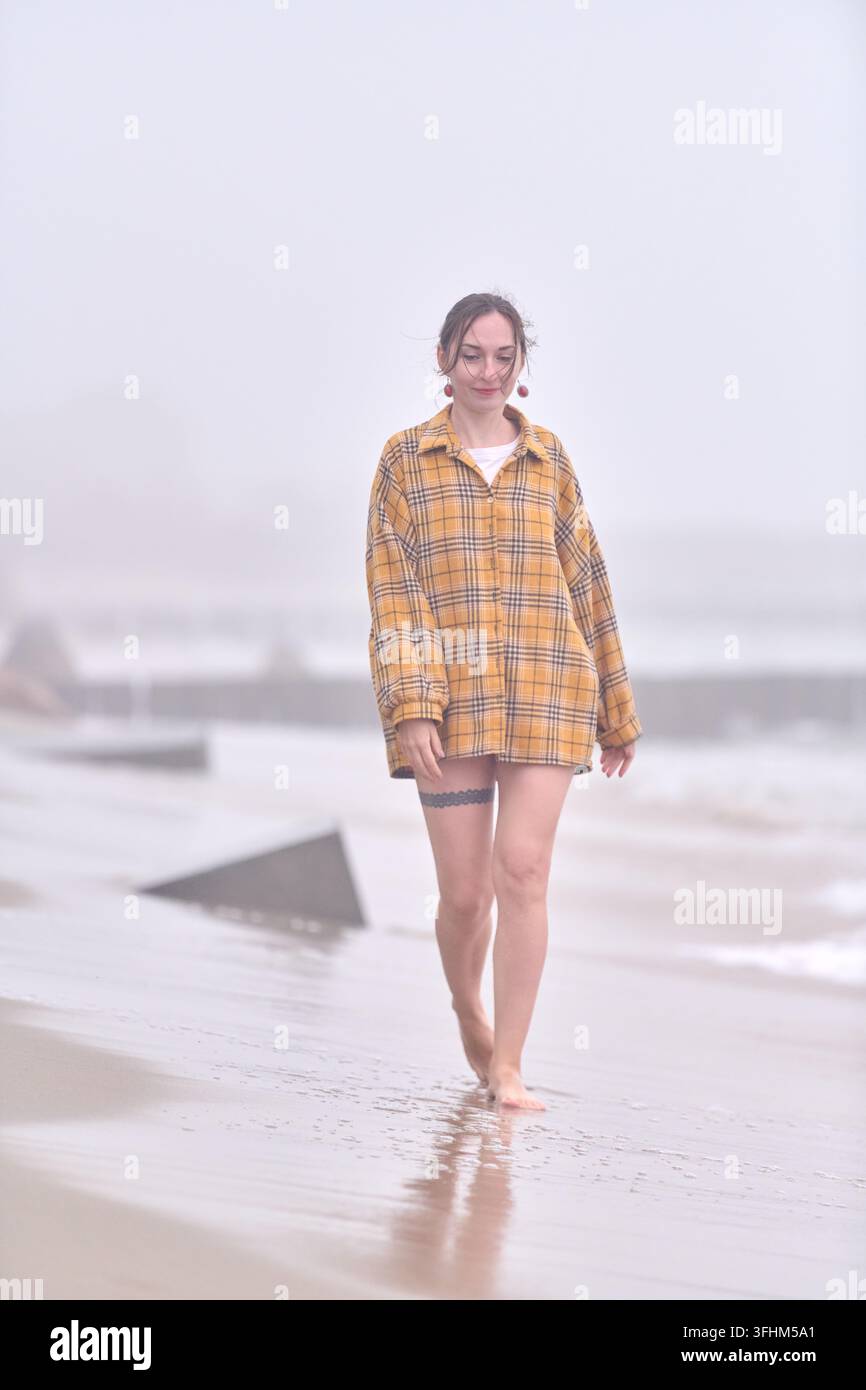 Giovane donna adulta con capelli castani che cammina a piedi nudi lungo una spiaggia nebbiosa in una camicia a quadri gialla, con occhi calmi, sabbia bagnata, schiuma pallida e struttura costiera sfocata Foto Stock