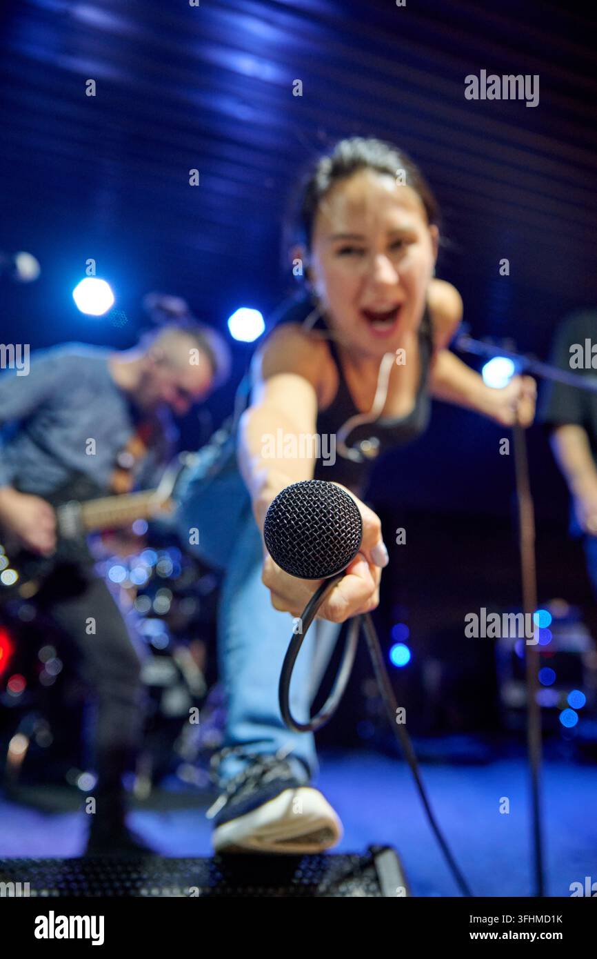 Giovane cantante con capelli scuri ed espressione intensa tende un microfono cablato verso la telecamera su un palco illuminato di blu accanto al chitarrista durante una performance dal vivo Foto Stock