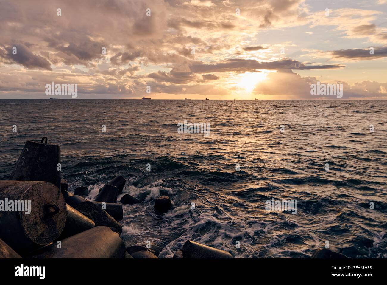 Onde oceaniche rotolanti circondano un frangiflutti di cemento frastagliato sotto un tramonto luminoso, con navi lontane lungo l'orizzonte e nuvole ramate sopra un mare texture Foto Stock