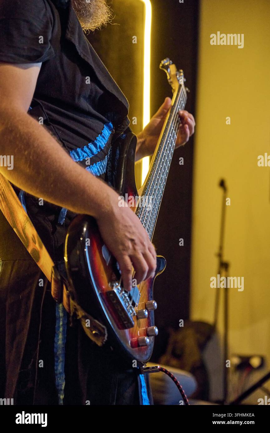 Musicista barbuto suona un basso elettrico lucido accanto a un supporto microfonico sotto una calda luce dorata del palco, con abiti scuri, accenti blu e sfondo sfocato Foto Stock
