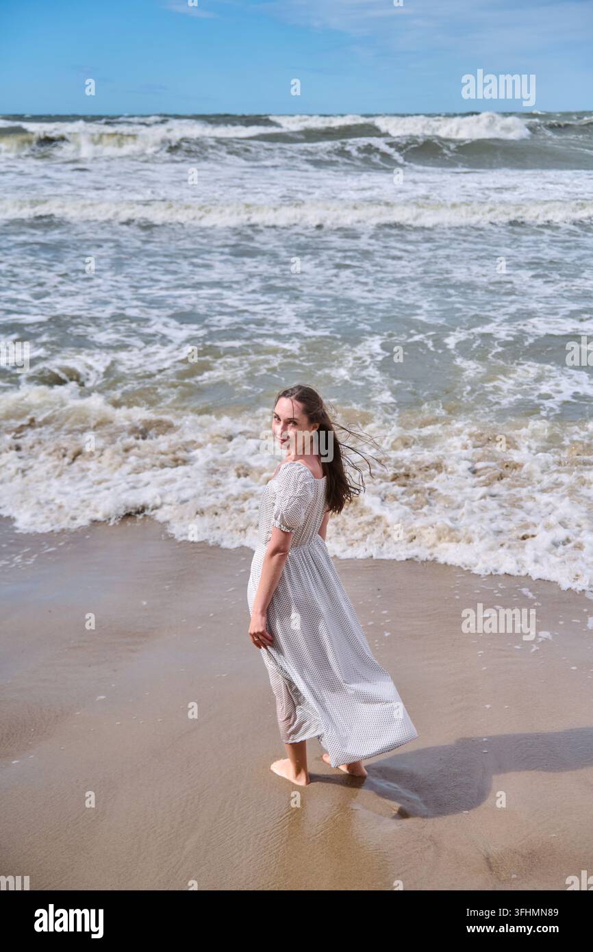 Donna in abito bianco fluente che cammina a piedi nudi lungo la spiaggia di sabbia, onde schiumose dietro, luce intensa, cielo blu, libertà costiera e tranquillo estate li Foto Stock