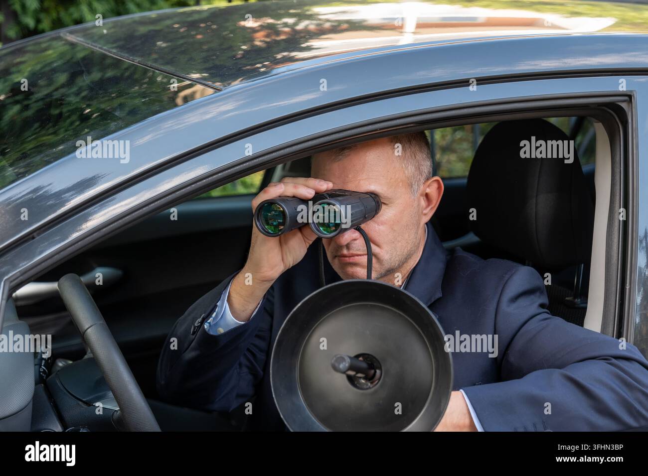 Investigatore privato monitora un obiettivo dalla sua auto con un binocolo mentre utilizza apparecchiature per l'ascolto remoto Foto Stock