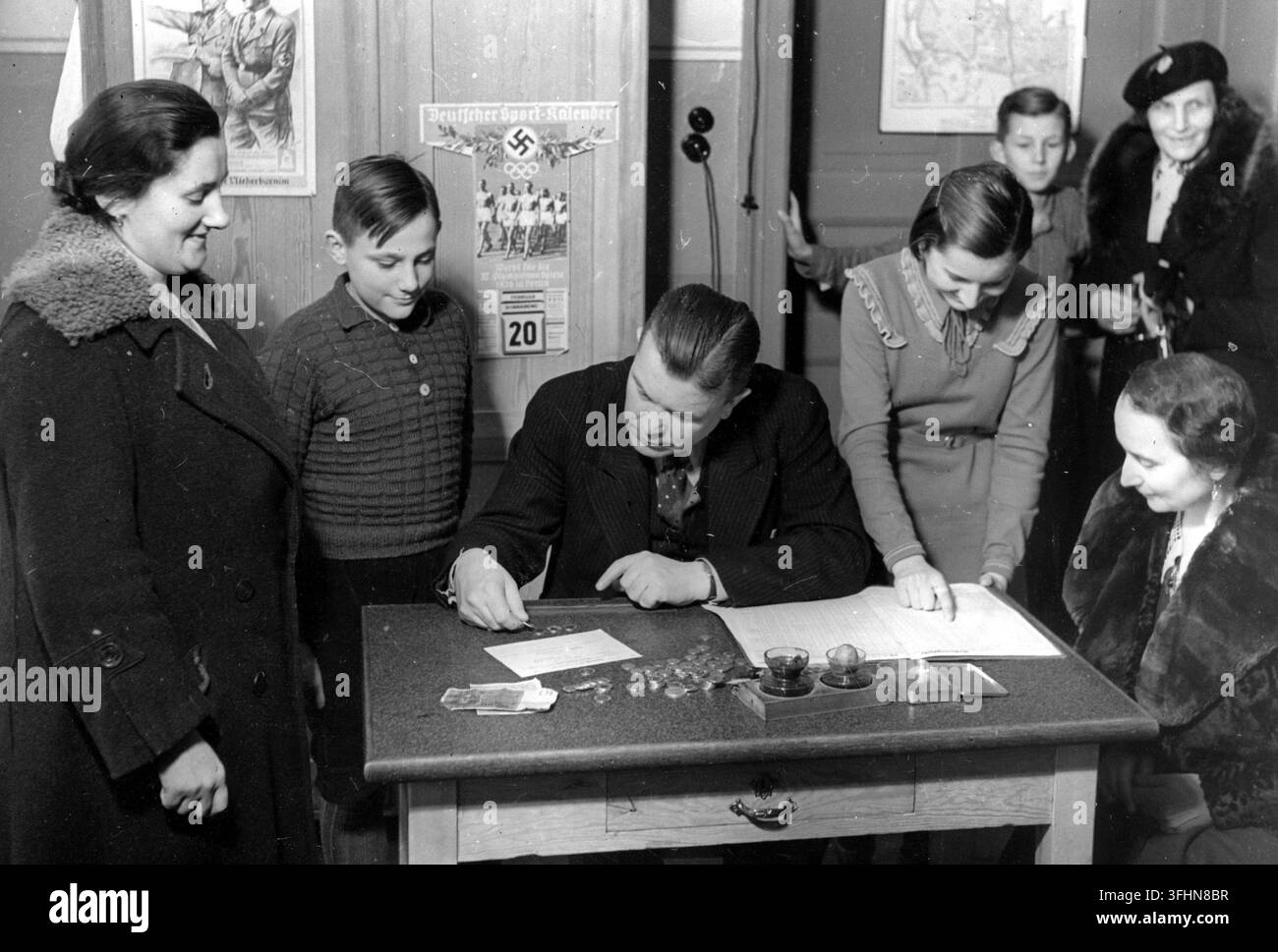 Un evento di raccolta di risparmi, 30 ottobre 1940, con un gruppo di adulti e bambini riuniti intorno a un tavolo di legno, dove un uomo in abito scuro conta monete e maneggia carte, in una stanza con poster e una mappa sulle pareti. Foto Stock