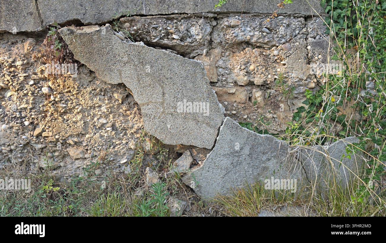 Grande sezione di un muro di sostegno in cemento rotto e trascurato che si sgretola nel terreno, rivelando aggregati di pietra interni con edera comune che cresce Foto Stock