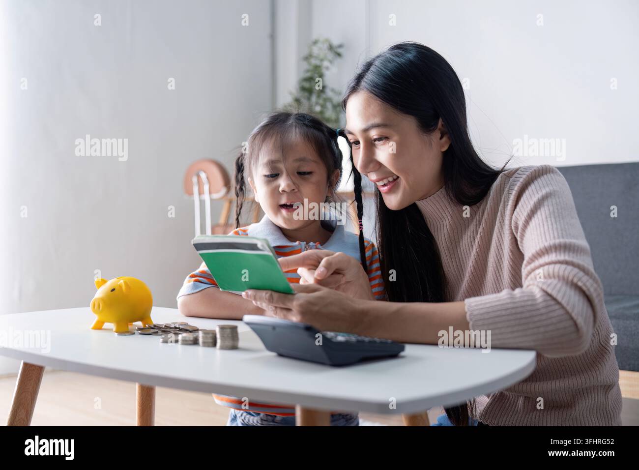 Madre e figlia diverse che imparano a risparmiare insieme con un maialino di gomma giallo, una calcolatrice e un quaderno a casa, enh Foto Stock