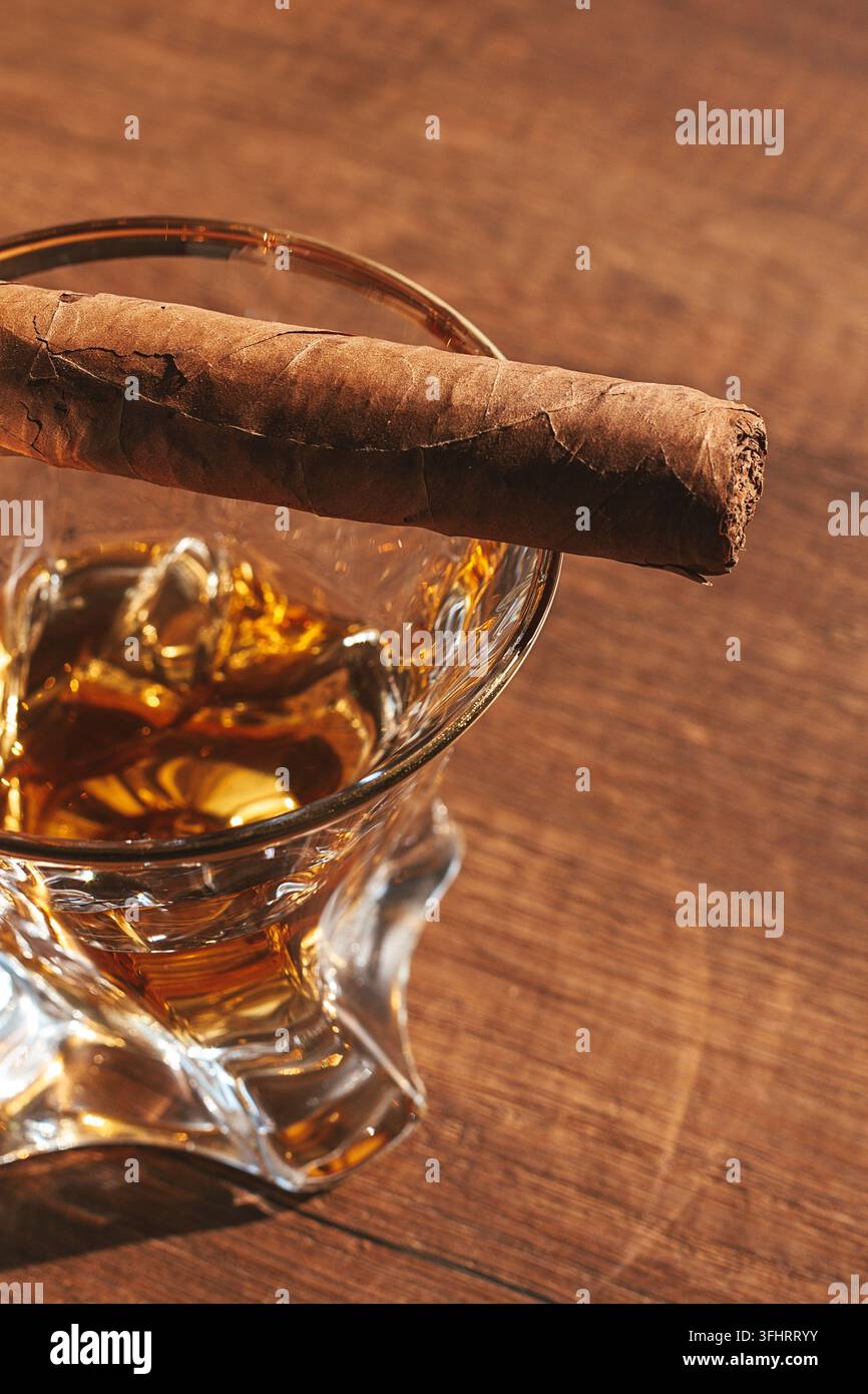 Un sigaro è posizionato sul bordo di un bicchiere pieno di whisky. L'ambientazione è un tavolo di legno, che suggerisce un'atmosfera informale. Questa scena cattura una mamma Foto Stock