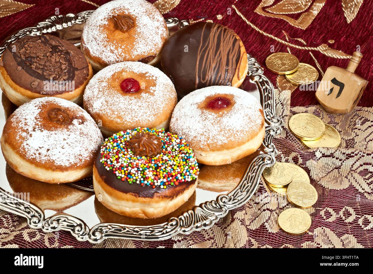 Ciambelle tradizionali per Hanukkah con gelatina, cioccolato e zuccherini su un vassoio d'argento accanto a un dreidel di legno e monete d'oro su una tovaglia rossa. Foto Stock