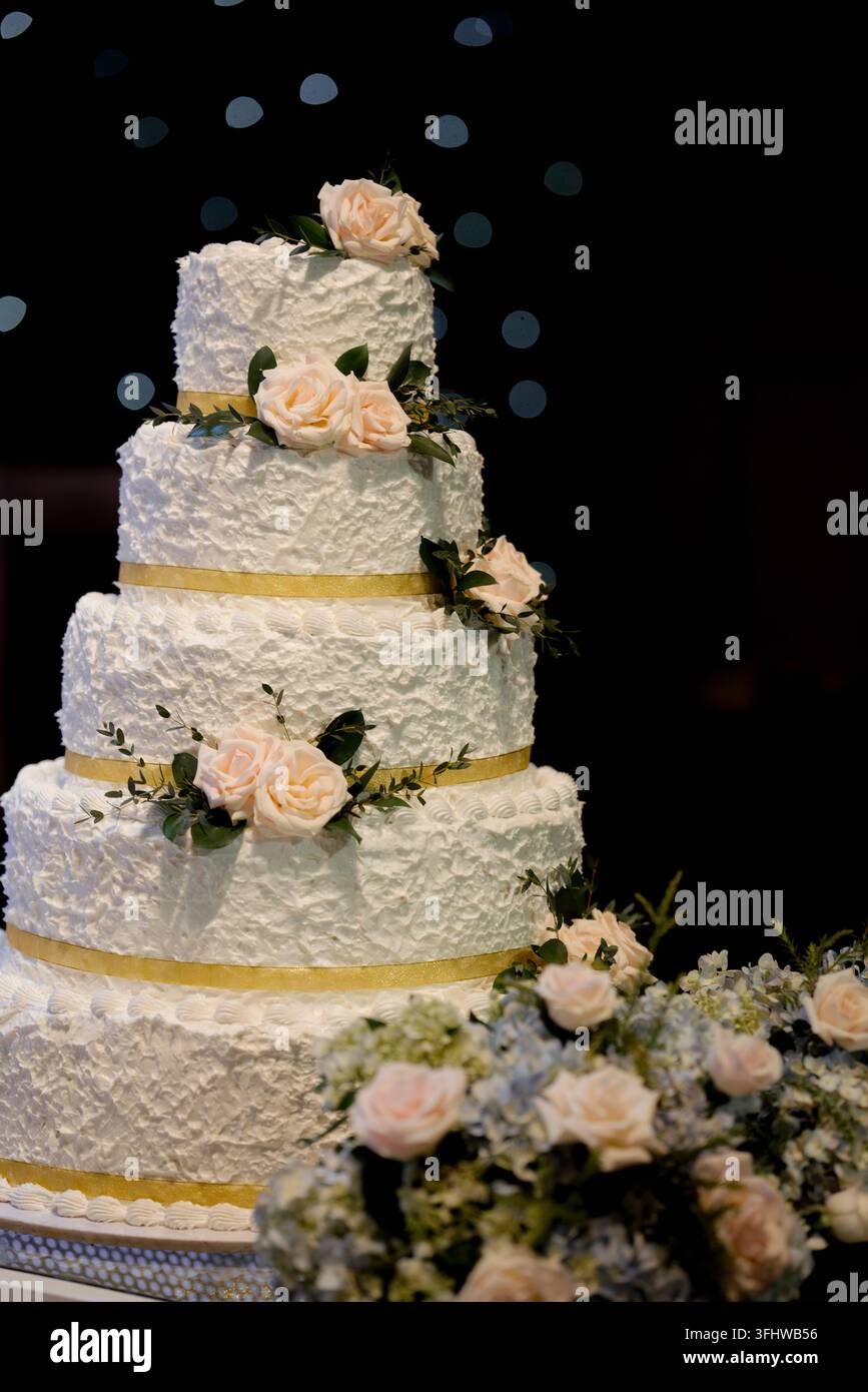 Elegante torta nuziale bianca a più piani decorata con rose pesche e nastro dorato Foto Stock