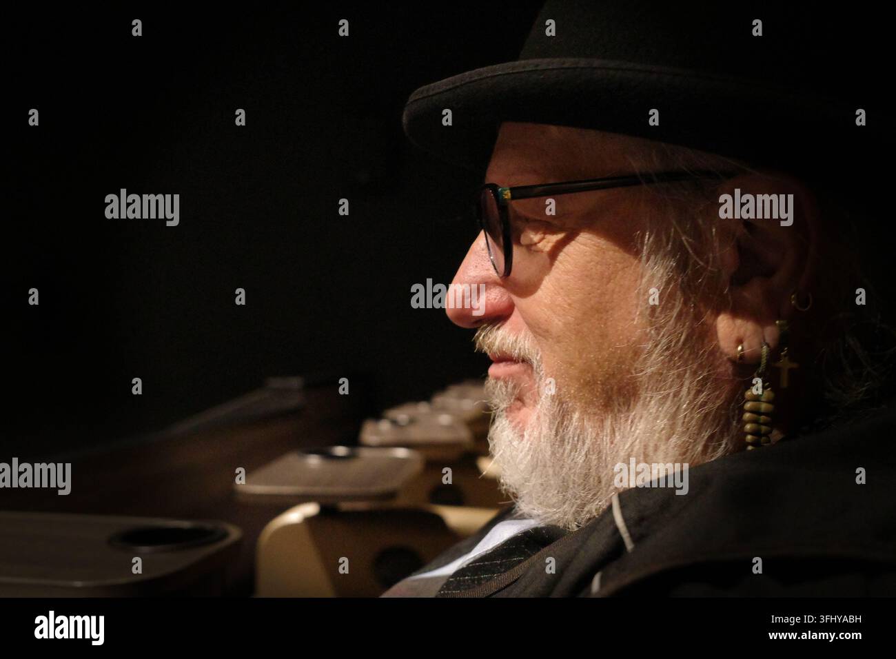 Un uomo anziano è seduto in un cinema. Indossa un cappello nero e occhiali, con un distinto apparato facciale che include baffi e barba. Il suo orecchio mostra più Foto Stock