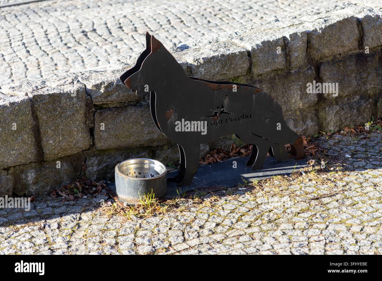 Szklarska Poręba, Polonia – 17 dicembre 2025: Scultura di un cane in metallo con una ciotola per bere su un marciapiede di pietra a Szklarska Poręba, che crea una comodità Foto Stock
