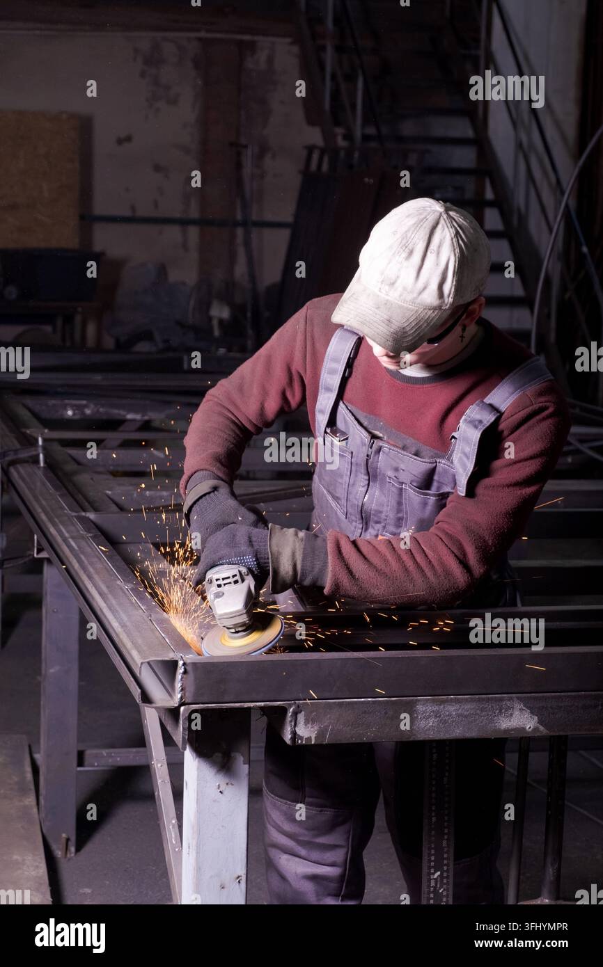 L'uomo che indossa il cappuccio e la tuta crea scintille durante la lucidatura dell'acciaio in officina Foto Stock