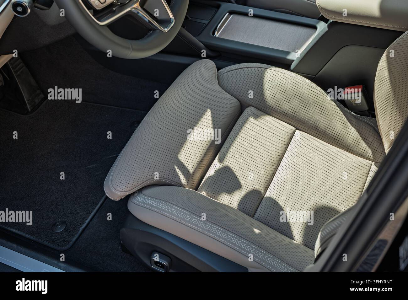 Colpo ad angolo alto di un sedile anteriore in pelle perforata beige chiaro, del bordo del volante e della console centrale di un'auto moderna parcheggiata all'aperto alla luce del giorno. Foto Stock