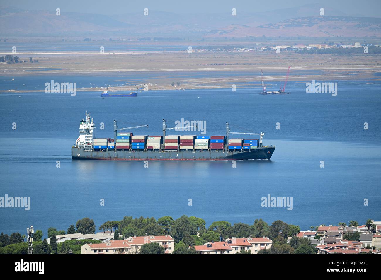 Izmir, Turchia – 18 settembre 2026: Una nave portacontainer carica di container di spedizione naviga lungo una baia costiera vicino agli edifici residenziali Foto Stock