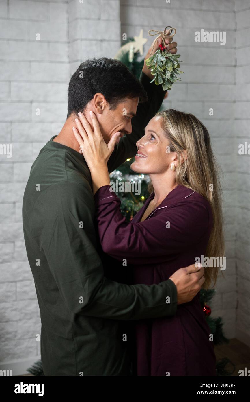 Coppia felice che condivide un momento romantico natalizio abbracciandosi sotto il vischio davanti a un albero di Natale decorato a casa. Foto Stock