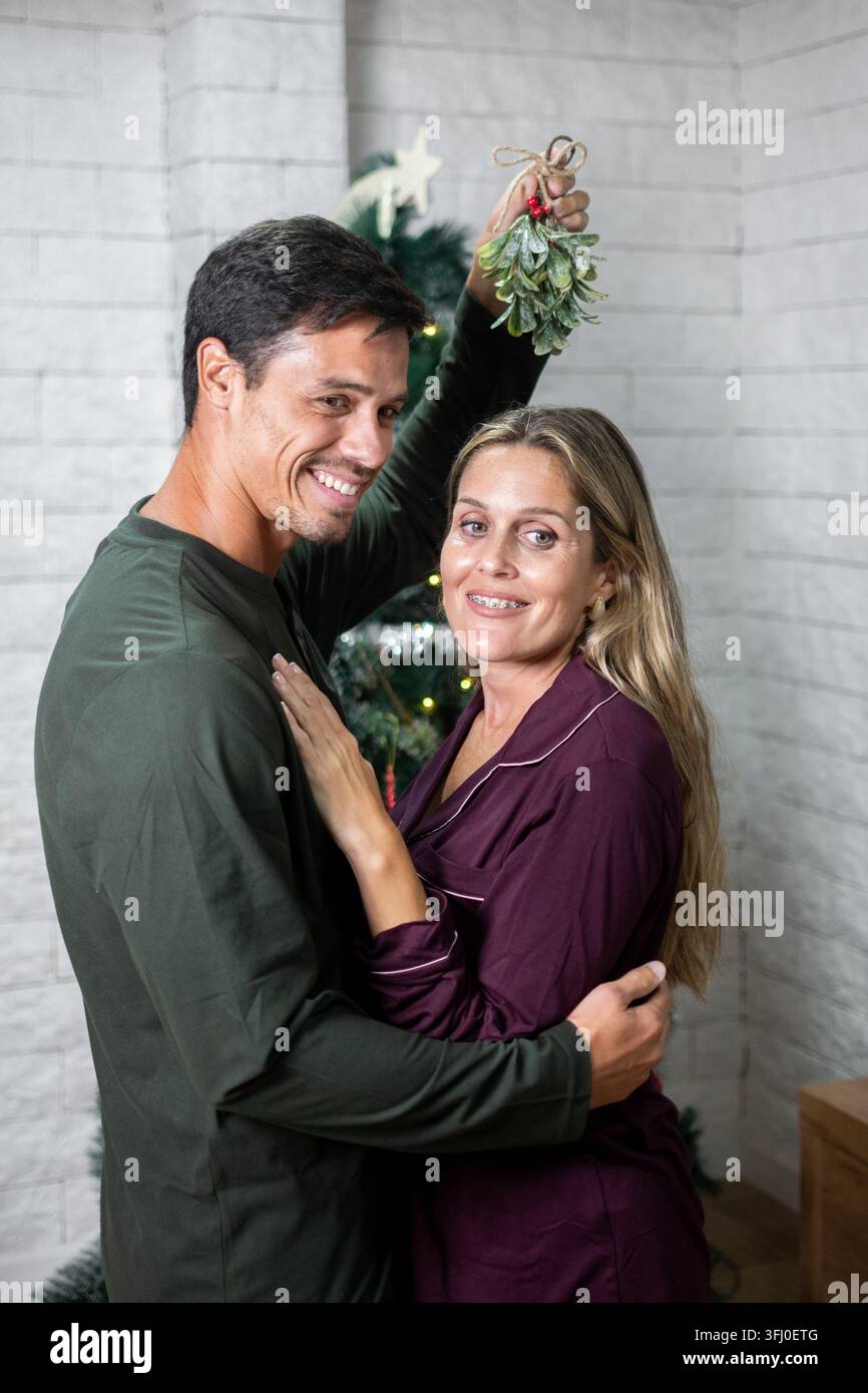 Coppia felice che condivide un momento romantico natalizio abbracciandosi sotto il vischio davanti a un albero di Natale decorato a casa. Foto Stock