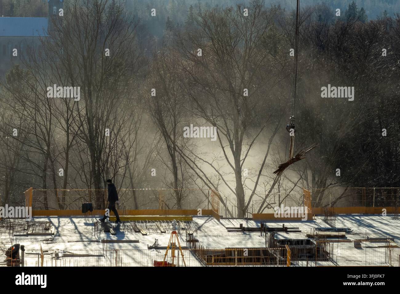 Un operaio industriale si muove lungo una struttura in cemento durante le ore di lavoro mattutine, mentre la luce dorata illumina la polvere atmosferica, simboleggiando la professione Foto Stock