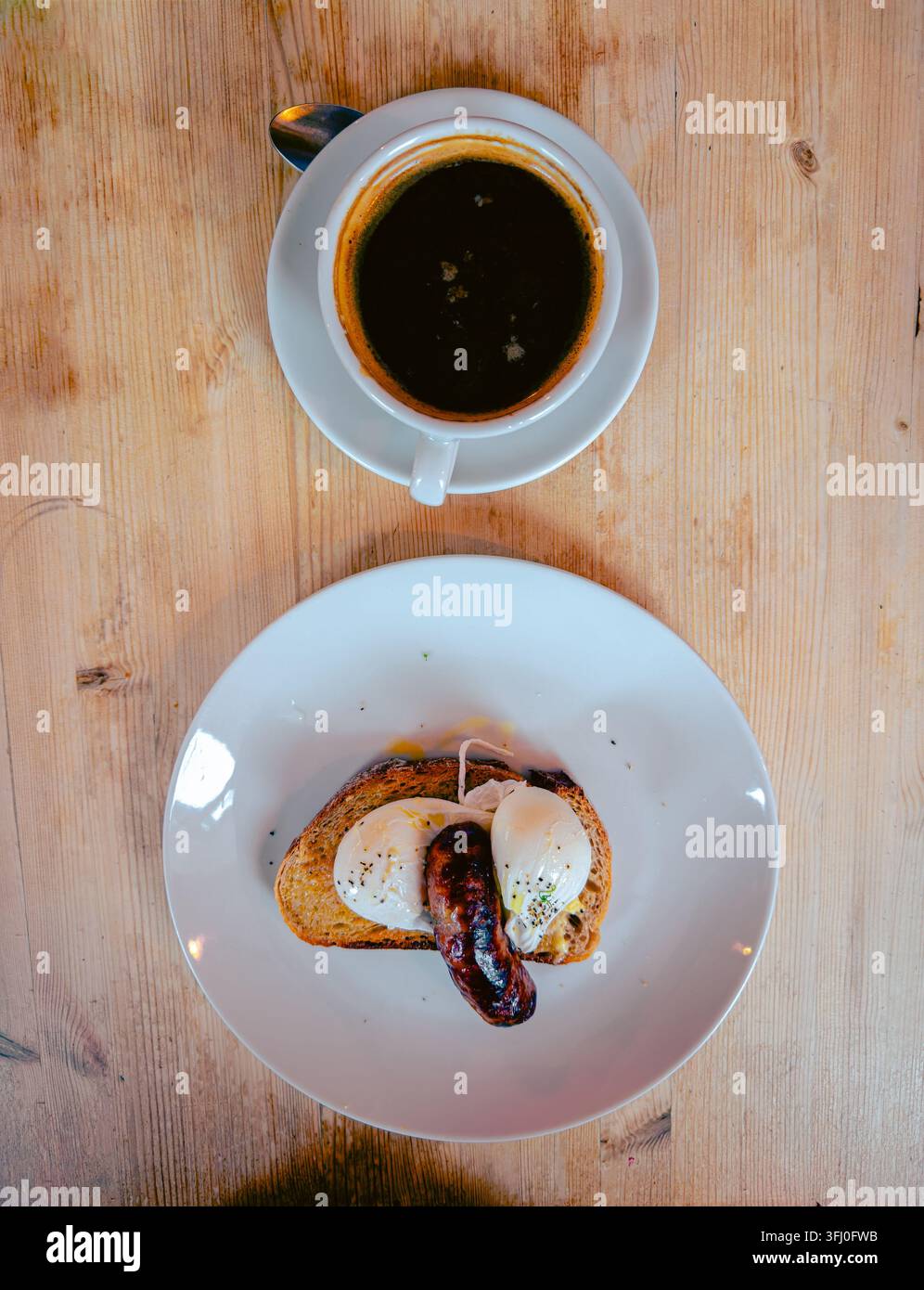 Vista dall'alto di un piatto da colazione con uova sode e una salsiccia grigliata sul toast, servito con una tazza di caffè nero su un tavolo di legno. - Immagine stock catturata con smartphone