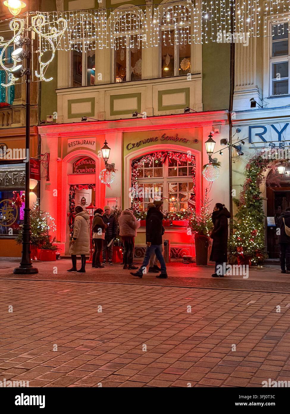 Pozna, Polonia – 30 dicembre 2025: Pedoni passeggiano davanti a un ristorante luminosamente illuminato durante la stagione delle feste invernali dove decorazioni stagionali luminose Foto Stock