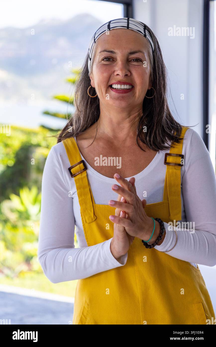 Donna di mezza età in piedi sulla porta e sorridente, indossa un grembiule giallo, una maglietta bianca, orecchini a cerchio. Terrazzo, giardino, esterno, illuminato dal sole Foto Stock
