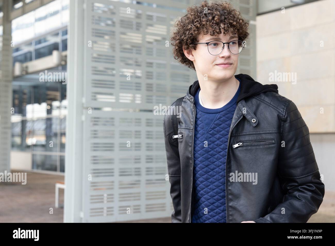 Ragazzo in piedi, che guarda di lato sulla piazza con giacca di pelle e cinghia dello zaino, spazio di copia. Urbano, pendolare, studente, occhiali Foto Stock