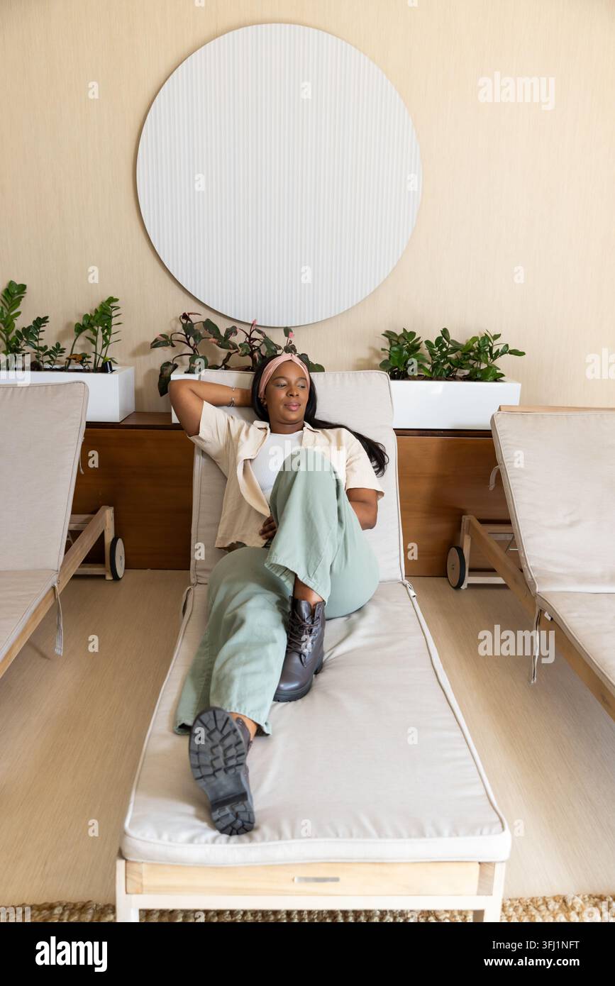 Donna africana sdraiata su una chaise longue in legno imbottita nella sala spa con vaso, tappeto, specchio Foto Stock